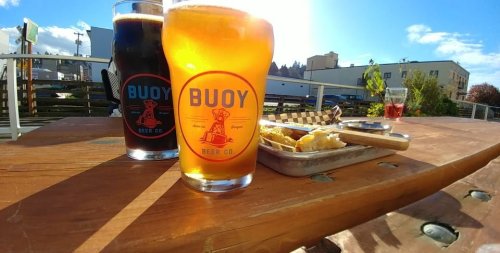 Two glasses of beer on a wooden table, with a plate of food and a sunny outdoor setting in the background.