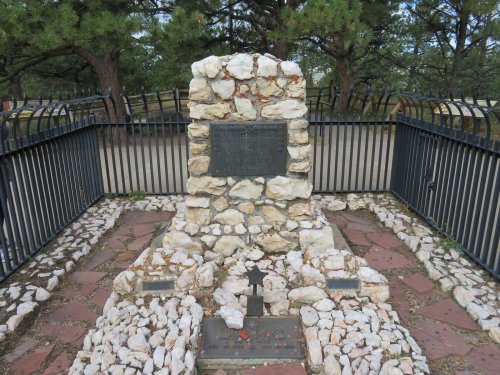 A stone monument surrounded by a black fence, with plaques and decorative stones in a wooded area.