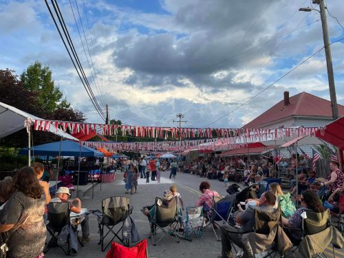A lively street festival with people sitting in chairs under colorful banners and tents, set against a cloudy sky.