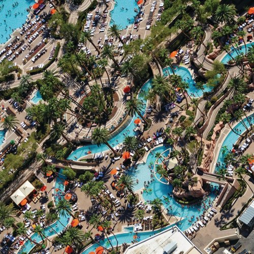Aerial view of a resort pool area with palm trees, lounge chairs, and winding water features.