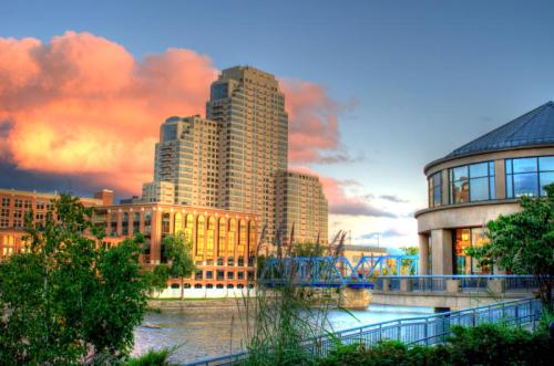 City skyline at sunset with tall buildings, a river, and a bridge, surrounded by greenery and colorful clouds.