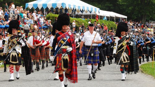 A parade of bagpipers and drummers in traditional Scottish attire, performing in front of a large crowd under a tent.