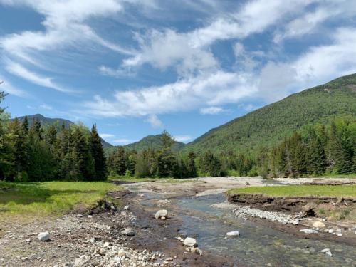 A serene landscape featuring a winding stream, lush green trees, and mountains under a blue sky with scattered clouds.
