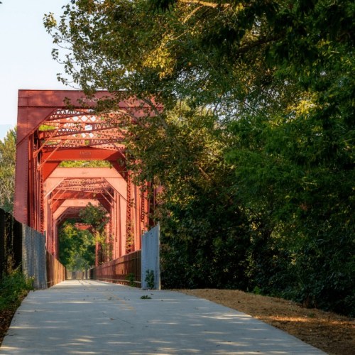 A red metal bridge spans a pathway surrounded by lush greenery, leading into the distance under clear blue skies.