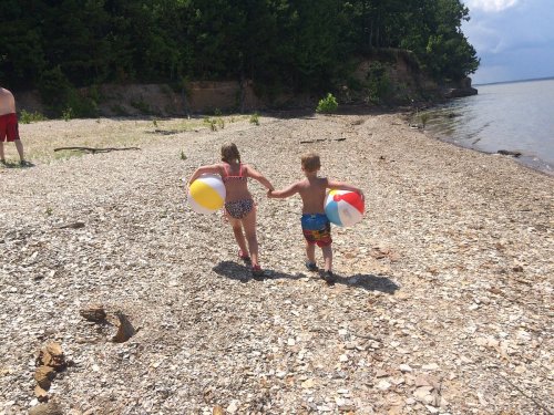 Two children holding hands, running along a rocky beach with beach balls, surrounded by trees and water.