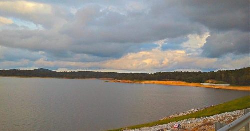 A serene lake surrounded by hills under a cloudy sky, with a sandy shore and rocky bank.