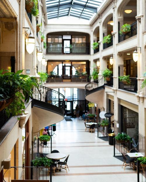 A spacious indoor atrium with plants, balconies, and seating areas, featuring natural light from a glass roof.