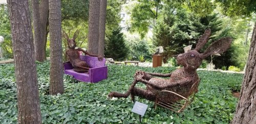Two large rabbit sculptures made of twigs sit in a green, leafy garden, surrounded by trees and other art pieces.