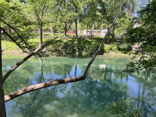 A serene river scene with clear blue water, surrounded by lush green trees and a grassy bank.