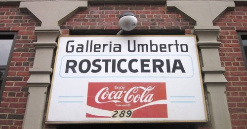 Sign for "Galleria Umberto Rosticceria" with a Coca-Cola logo, mounted on a brick wall.
