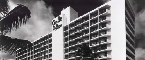 Black and white image of a modern hotel building with balconies, surrounded by palm trees and a cloudy sky.