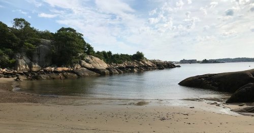 A serene beach scene with rocky shores, calm water, and lush greenery under a partly cloudy sky.