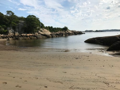 A serene beach scene with calm water, rocky shores, and trees under a partly cloudy sky.