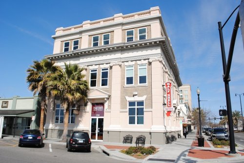 Historic building with palm trees, featuring large windows and a red sign, located on a sunny street corner.