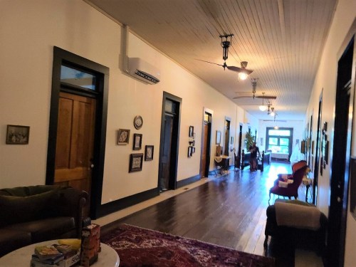 A long hallway with wooden floors, framed pictures on the walls, and vintage furniture, leading to a bright room at the end.