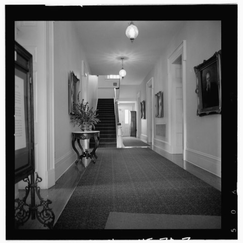 A black and white photo of a hallway with a staircase, framed portraits, and decorative lighting.