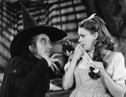 A witch in a black hat menaces a girl in a gingham dress, both in a dramatic black-and-white scene.