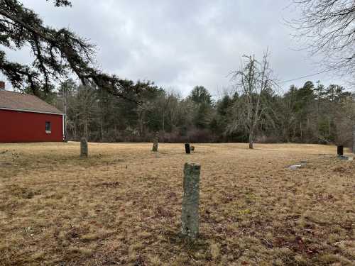 A grassy field with several stone markers and a red building in the background, surrounded by trees under a cloudy sky.