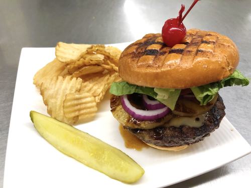 A grilled burger topped with lettuce, onion, pineapple, and cheese, served with chips and a pickle on a white plate.