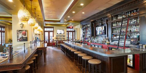 A stylish bar interior with wooden stools, a long counter, and shelves filled with various bottles. Warm lighting enhances the ambiance.