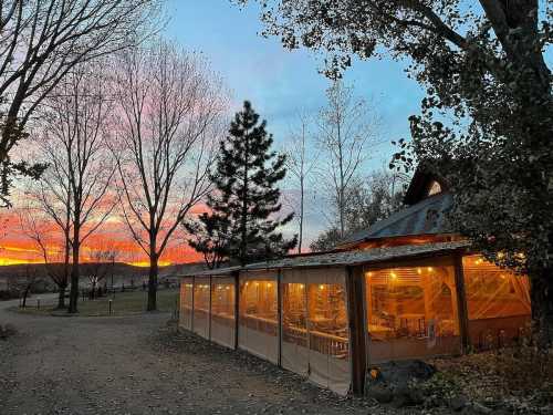 A cozy outdoor dining area with warm lights, surrounded by trees, under a colorful sunset sky.