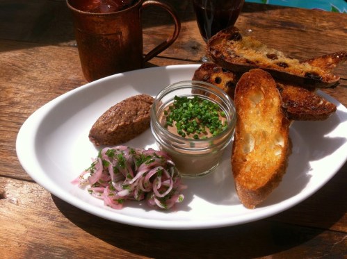 A plate with toasted bread, a jar of pâté topped with chives, and a side of pickled onions. A drink is in the background.