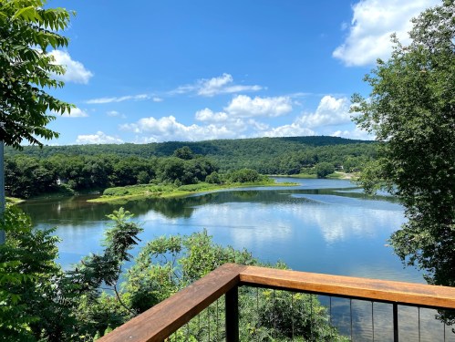 A serene lake surrounded by lush greenery and hills under a bright blue sky with fluffy white clouds.