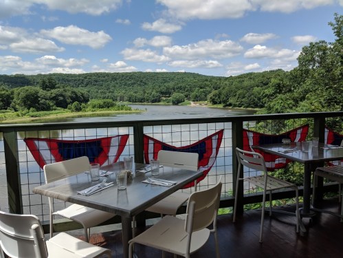 A scenic view from a restaurant deck overlooking a river, with mountains in the background and flags hanging.