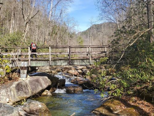 A hiker crosses a wooden bridge over a stream, surrounded by trees and rocky terrain in a serene natural setting.