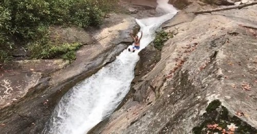 A person slides down a natural rock water slide, surrounded by greenery and flowing water.