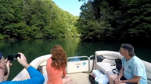 A group on a boat enjoying a scenic view of a calm, green river surrounded by lush trees.