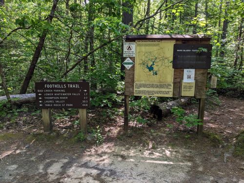 Trailhead sign for Foothills Trail, featuring a map and directions to nearby attractions in a lush, green forest.