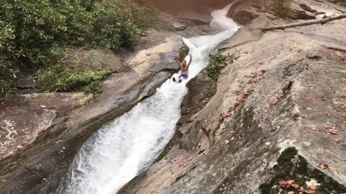 A person slides down a rocky water slide into a rushing stream surrounded by greenery.