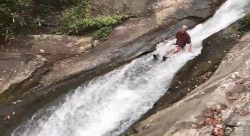 A person slides down a natural water slide, surrounded by rocks and greenery, with water flowing rapidly beside them.