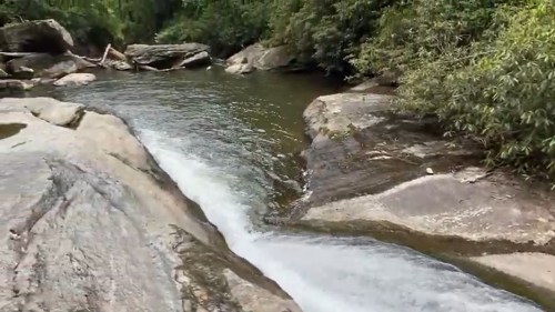 A serene river flows over smooth rocks, surrounded by lush greenery and trees.