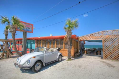 A vintage convertible parked outside a beachside restaurant with palm trees and a clear blue sky.