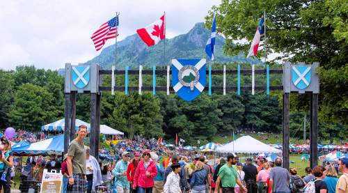 A lively festival scene with crowds under flags and a mountain backdrop, featuring tents and a festive atmosphere.