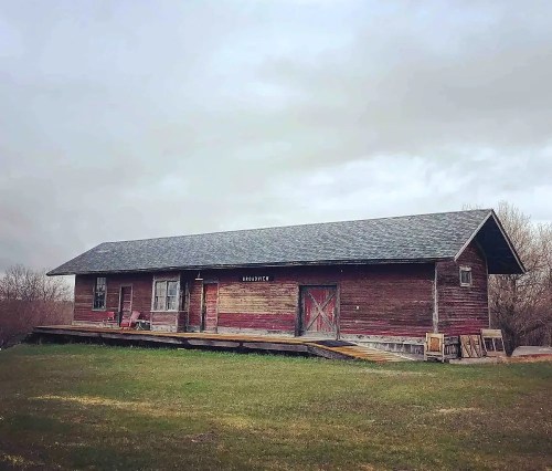 A rustic wooden house with a sloped roof, set on a grassy area under a cloudy sky.