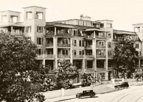 Historic building with multiple balconies, surrounded by trees and vintage cars parked in front.