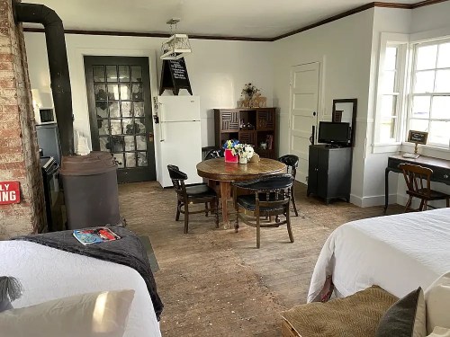 Cozy room with a dining table, chairs, a kitchenette, and large windows, featuring rustic decor and wooden floors.