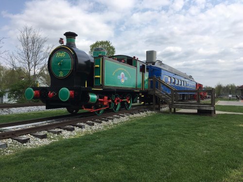 A vintage green steam locomotive on tracks, with a blue train car and grassy area in the background under a cloudy sky.