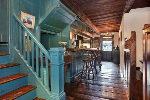 A cozy interior featuring a wooden bar, stools, and a staircase, with teal walls and rustic wooden beams.