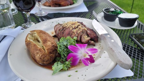 A plate with sliced steak, a baked potato, and a garnish of kale and an orchid flower, set on a table outdoors.