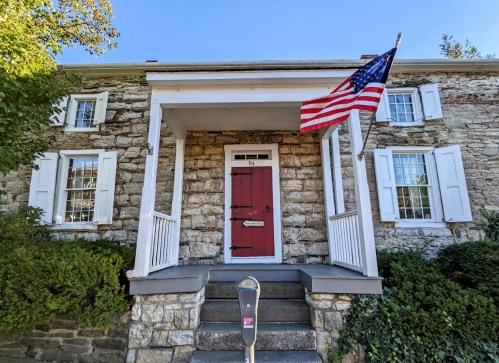 A stone house with a red door, white shutters, and an American flag on the porch, surrounded by greenery.