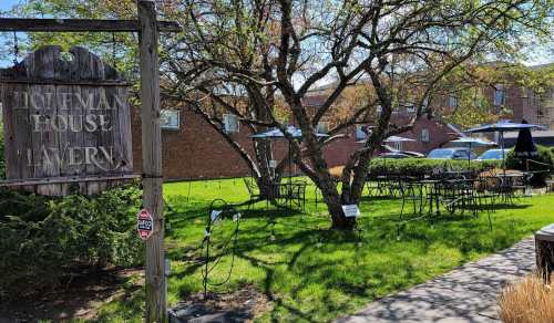 A sign for Hoffman House Tavern in a green outdoor area with tables, chairs, and trees on a sunny day.