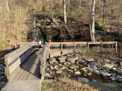 A wooden bridge leads to a small waterfall surrounded by trees and rocky terrain, with people enjoying the scenery.