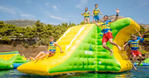 Children in life jackets jumping off a colorful inflatable water slide into a lake, surrounded by greenery.