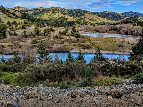 A scenic landscape featuring a winding river surrounded by lush greenery and rolling hills under a blue sky.