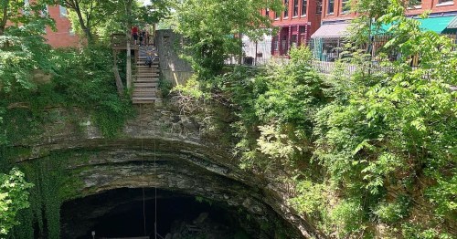 A deep, lush green sinkhole surrounded by trees, with a wooden platform and buildings in the background.