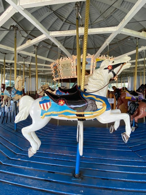 A white carousel horse with colorful decorations stands on a blue platform, surrounded by other horses in a vintage carousel.